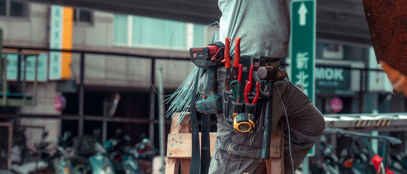 師傅繫著裝滿工具的腰帶站在梯子上施工，背景是街邊騎樓與機車。